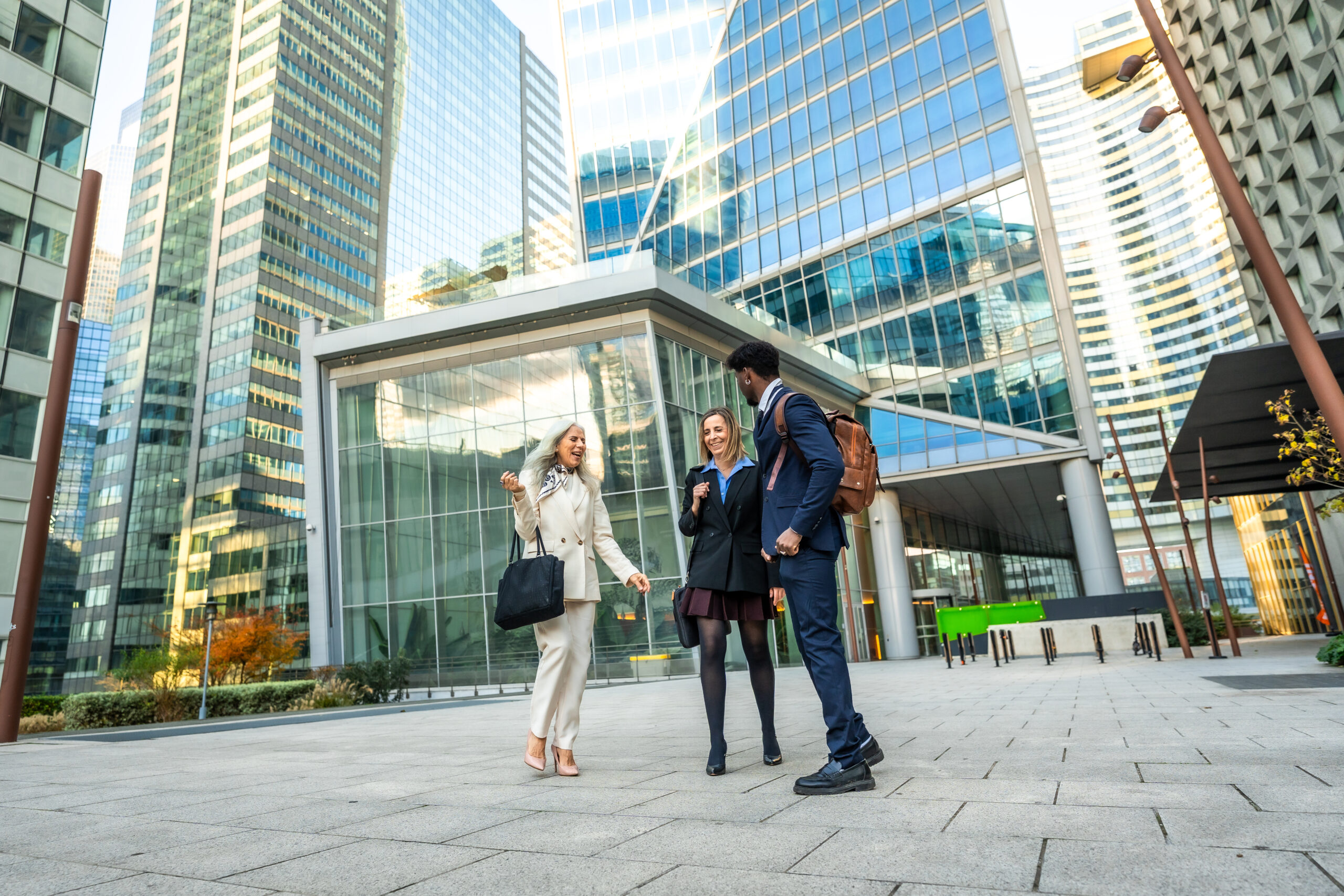 Diverse business colleagues engaging in cheerful conversation while walking through a modern urban district, surrounded by contemporary architecture and corporate skyscrapers on a bright day