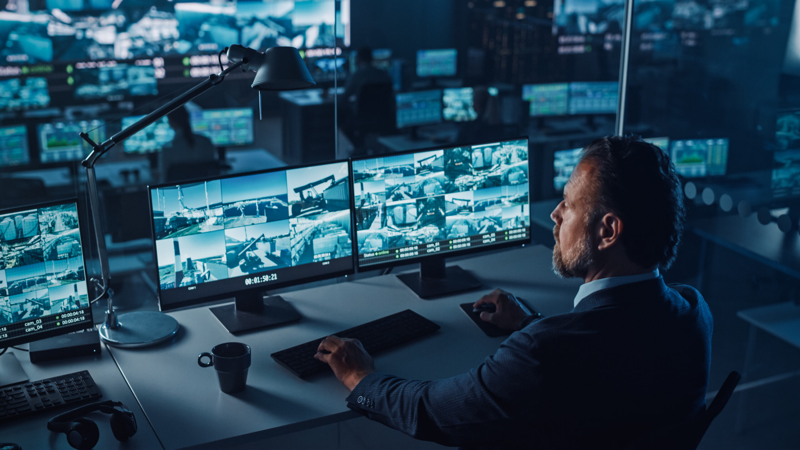 Male Officer Works on a Computer with Surveillance CCTV Video Footage in a Harbour Monitoring Center with Multiple Cameras on a Big Digital Screen. Employees Sit in Front of Displays with Big Data.